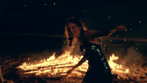 sparkly-green-dress-wearing young person with glitter on their face, hair in braids, dancing swimmingly in front of a roaring bonfire on a beach, mild ocean waves in the background coming toward her effortlessly